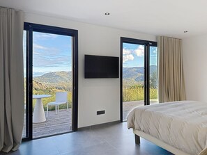 A bedroom with a bed, a TV, and a balcony with a table and chair. - Luxury villa, infinity pool, panoramic mountain view (Lumio)