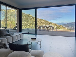 A modern living room with a sofa, a coffee table, and a dining area with chairs and a table, all with a view of a mountainous landscape. - Luxury villa, infinity pool, panoramic mountain view (Lumio)