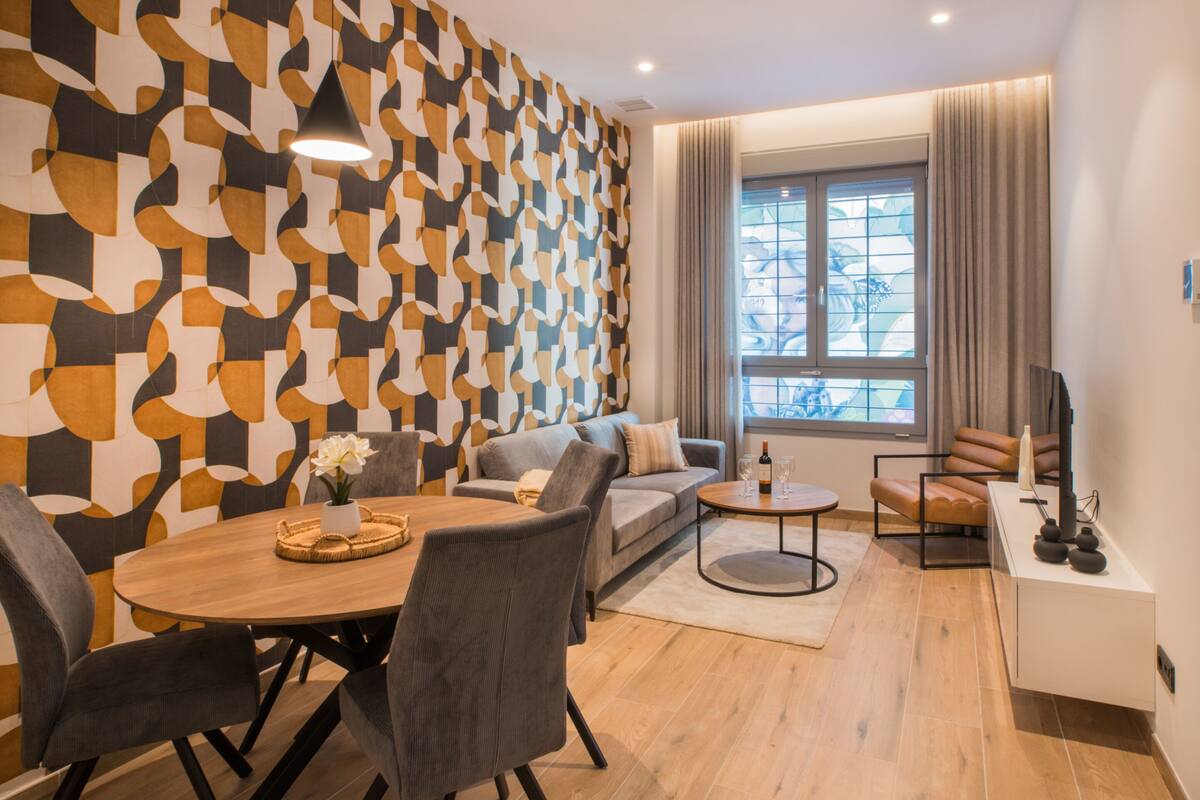 En moderne stue med væg i geometrisk mønster, sofa, spisebord med stole, sofabord og TV-møbel.