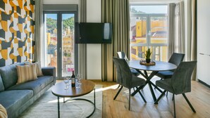 Un salon moderne comprenant un canapé, une table à manger avec des chaises, un téléviseur à écran plat fixé au mur et une grande fenêtre donnant sur des immeubles.