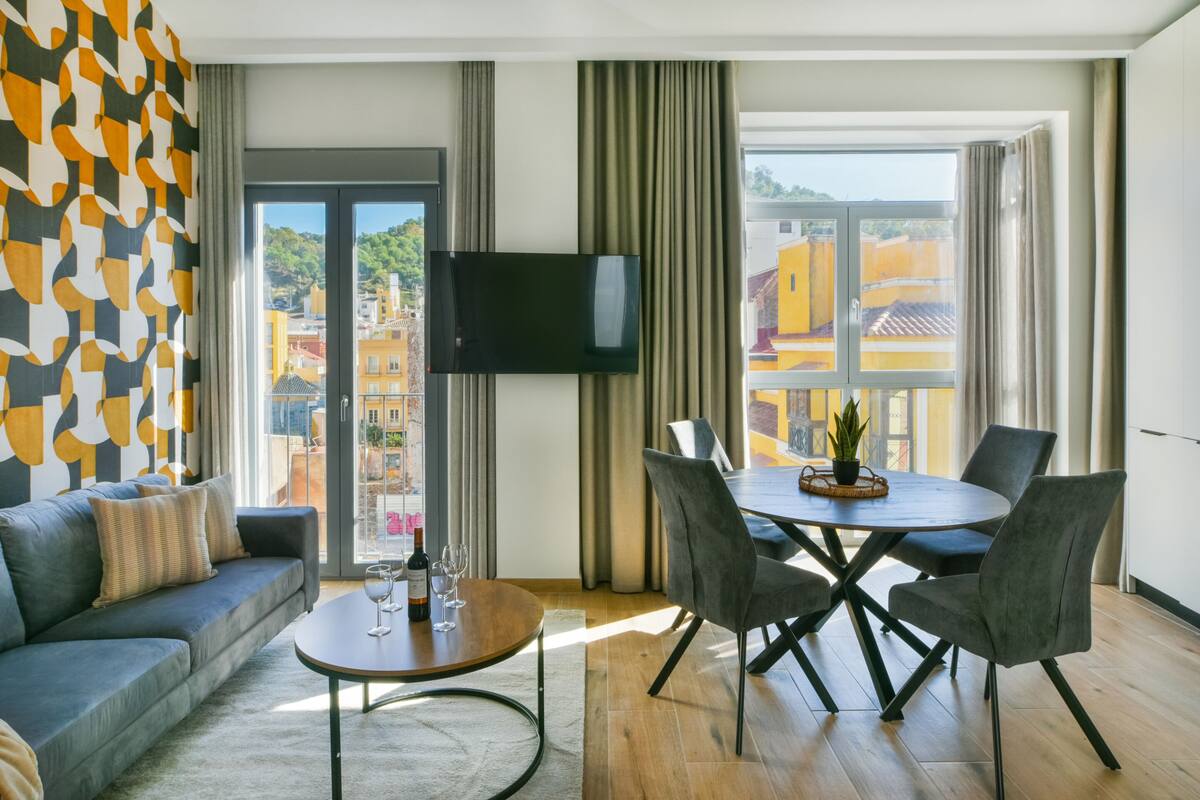 En moderne stue med sofa, spisebord med stole, et fladskærms-tv monteret på væggen og et stort vindue med udsigt til bygninger.