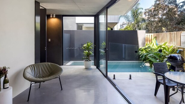 Un patio exterior moderno con puerta de vidrio que da a una piscina, una mesa de comedor con sillas y una planta en maceta.
