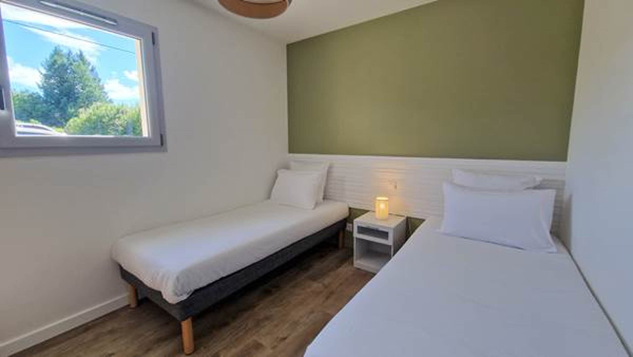 Een kleine kamer met een enkel bed, een raam, een nachtkastje met lamp en een groene accentmuur.