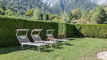 Ein Garten mit drei Liegestühlen und einem Tisch, umgeben von ordentlich geschnittenen Hecken und Blick auf die Berge.