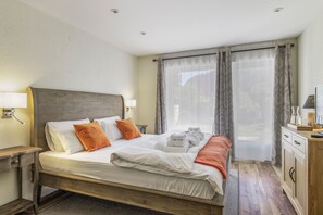 Une chambre d’hôtel avec un lit en bois, du linge de lit blanc, une table de chevet, un bureau avec une lampe et une vue sur l’extérieur à travers des rideaux légers.