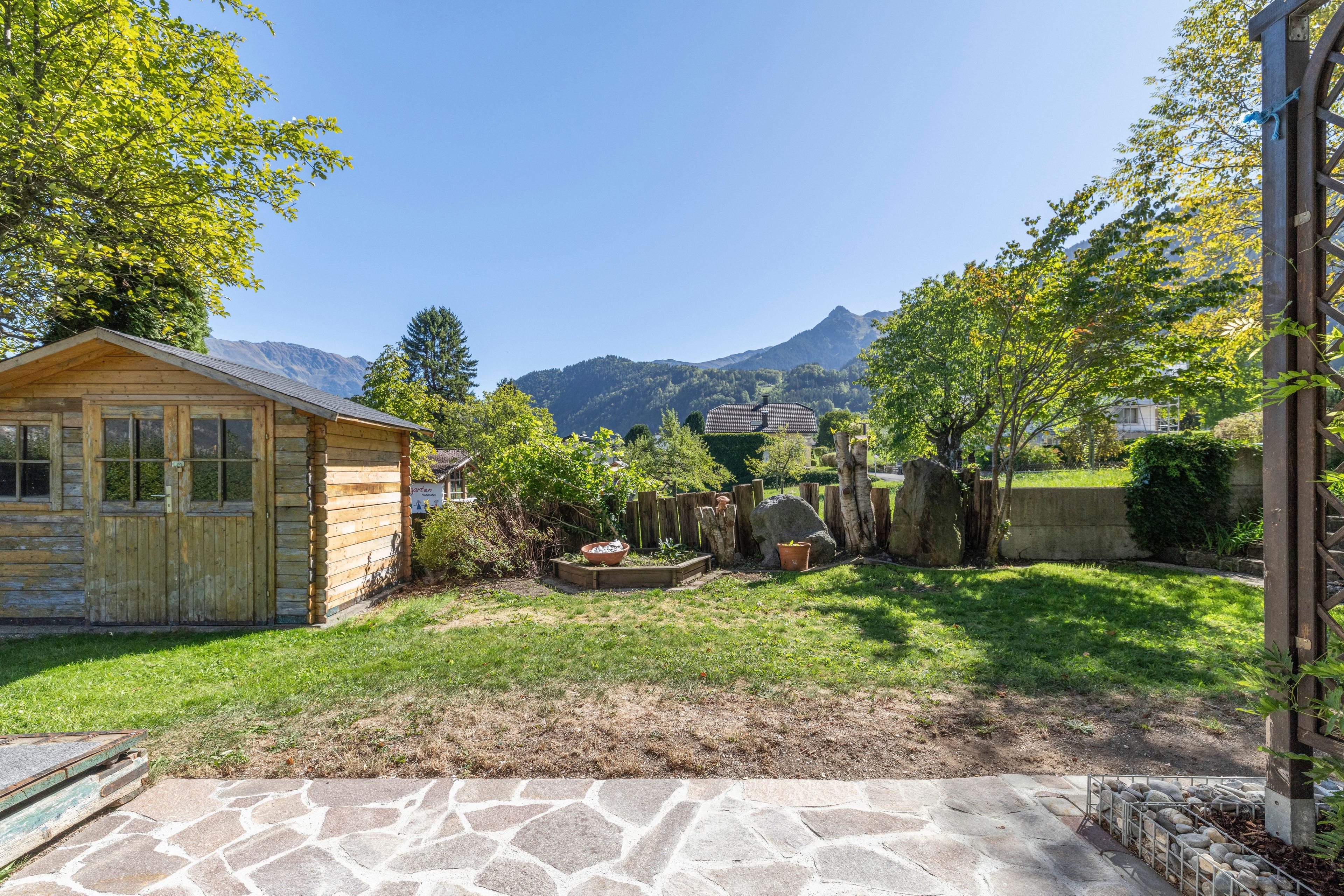 Eine Holzhütte mit einer Steinterrasse, umgeben von einem Garten und Blick auf die Berge.