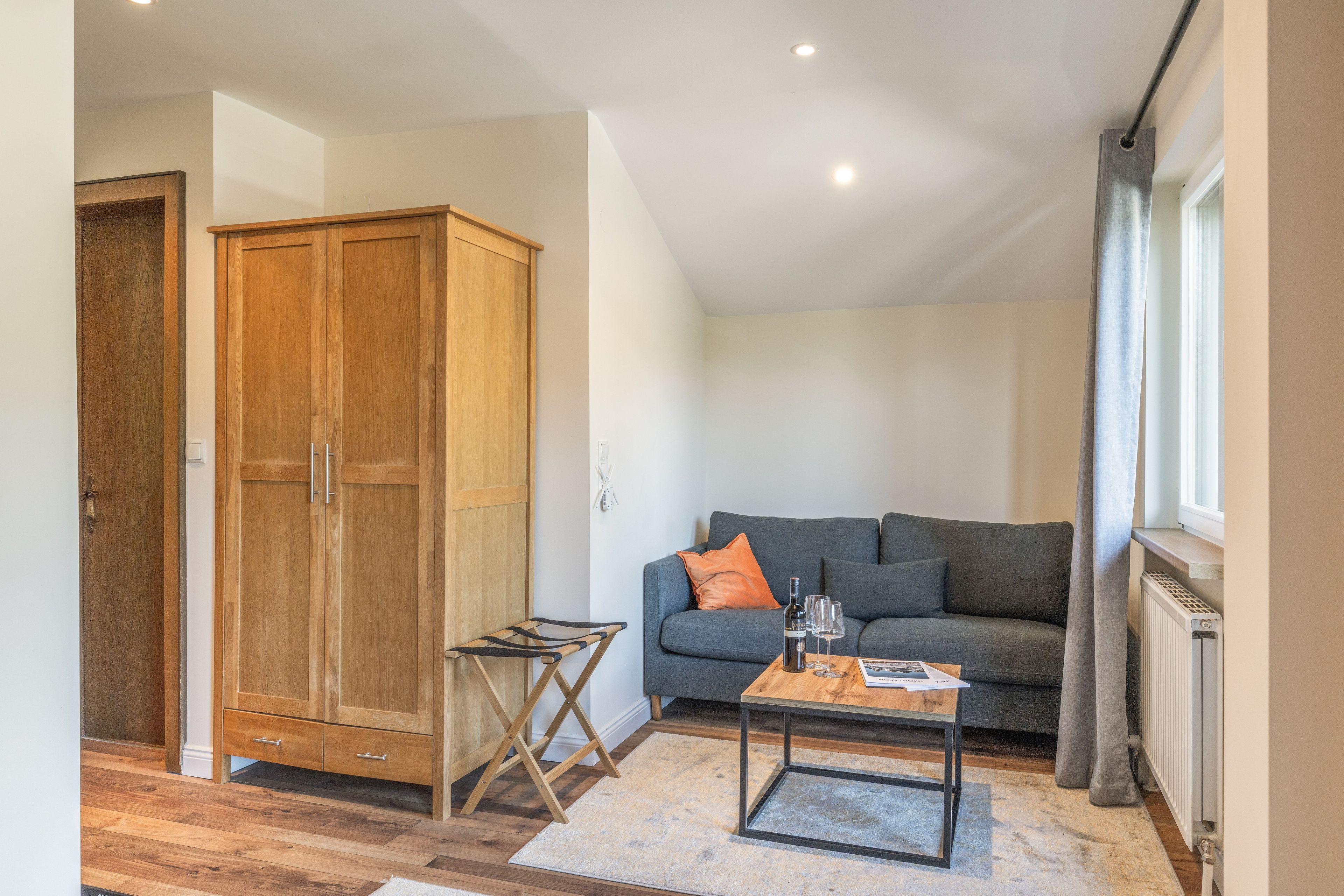 A room with a wooden wardrobe, a grey sofa, a small wooden table with a magazine, and a radiator.