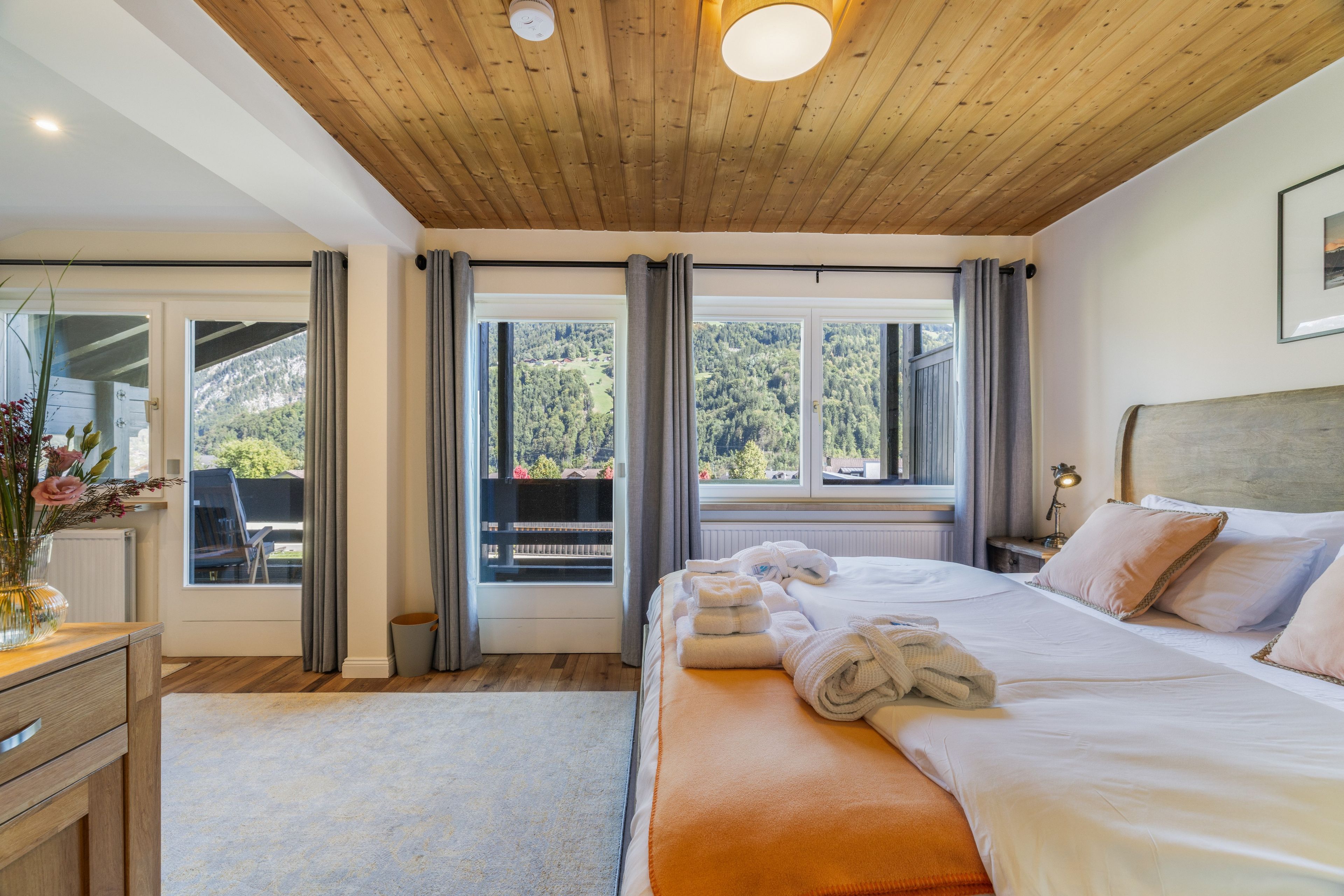A hotel room with a wooden ceiling, a bed with white bedding, a nightstand with a lamp, and a view of a forest through the window.