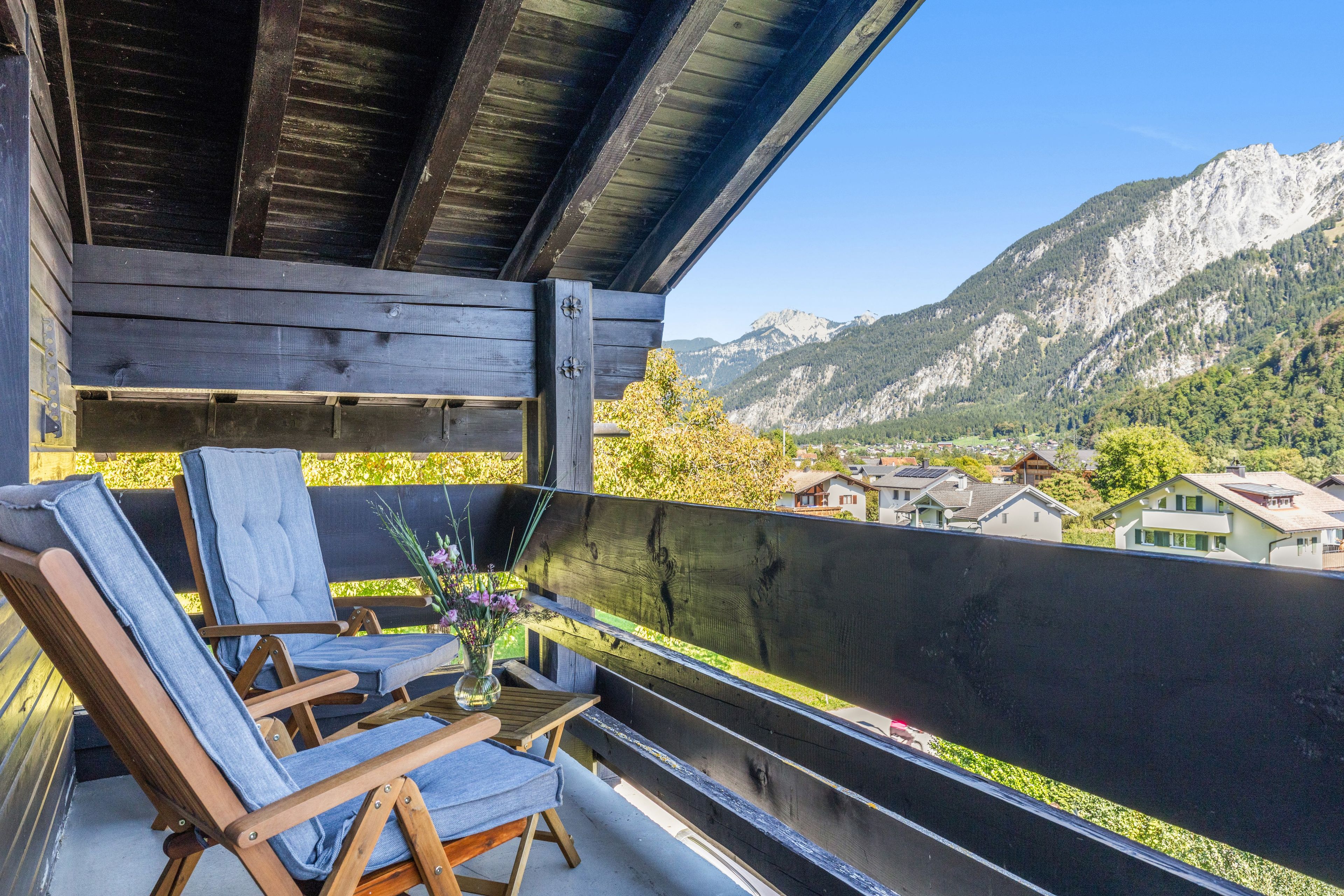 A balcony with wooden chairs and a table, overlooking a mountainous landscape with houses.