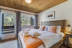 A bedroom with a large bed, a view of the outdoors, and a wooden ceiling.