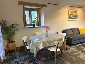 A dining area with a round table, chairs, a vase with flowers, a window with curtains, a sofa, and a painting on the wall. - Rest, Reconnect & Gather at a Historic Cornish Barn (Saint Clether)