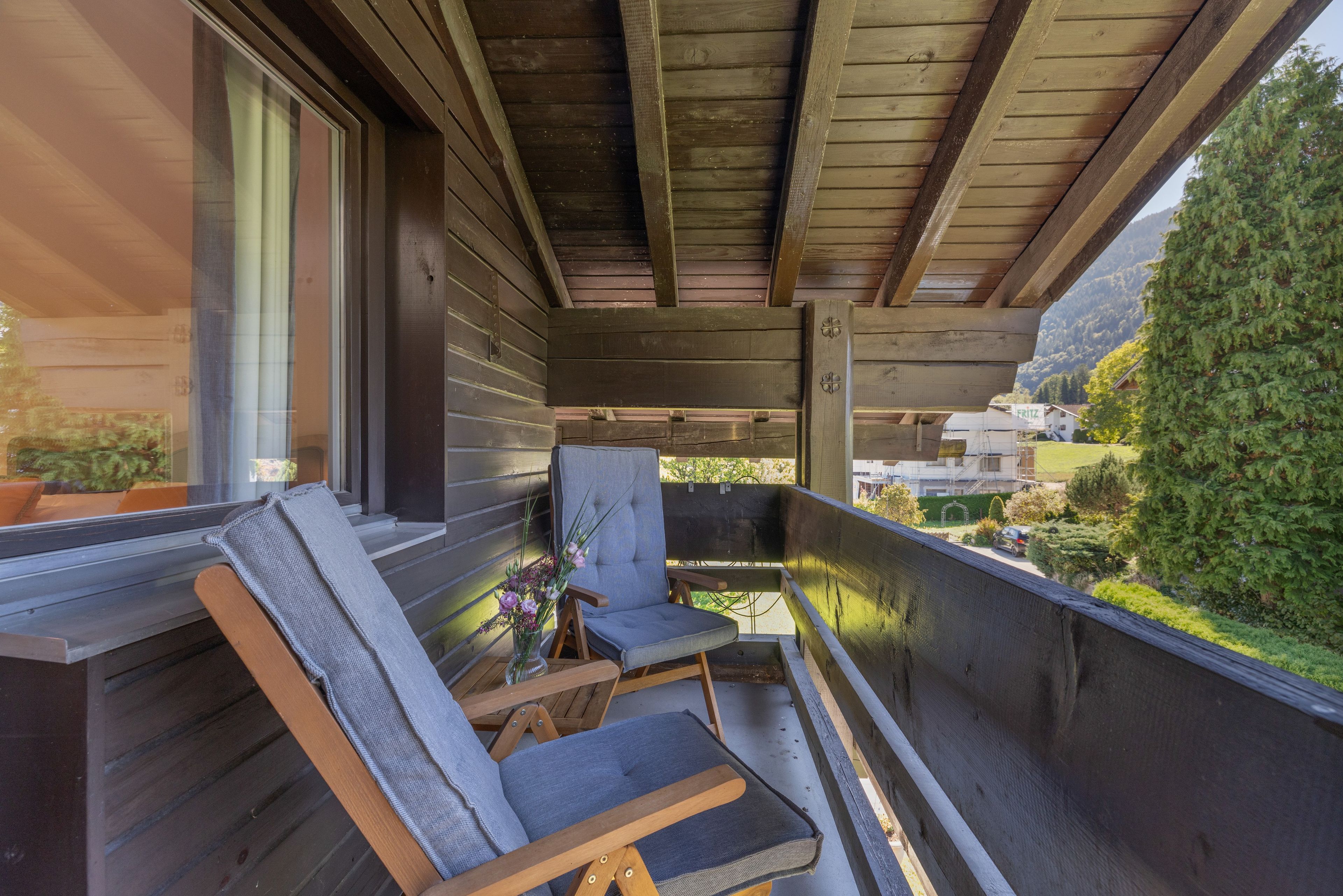 Ein Holzbalkon mit zwei Stühlen und einem Tisch, der auf einen Wohnbereich mit Häusern und Grünflächen blickt.
