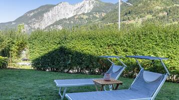 A pair of sun loungers with a table and a decorative vase, set on a grassy lawn with a mountain backdrop.