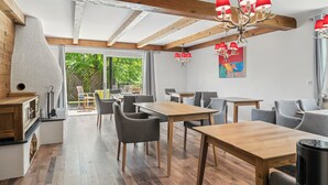 A modern dining area with wooden tables and chairs, a chandelier, and a view of greenery outside.