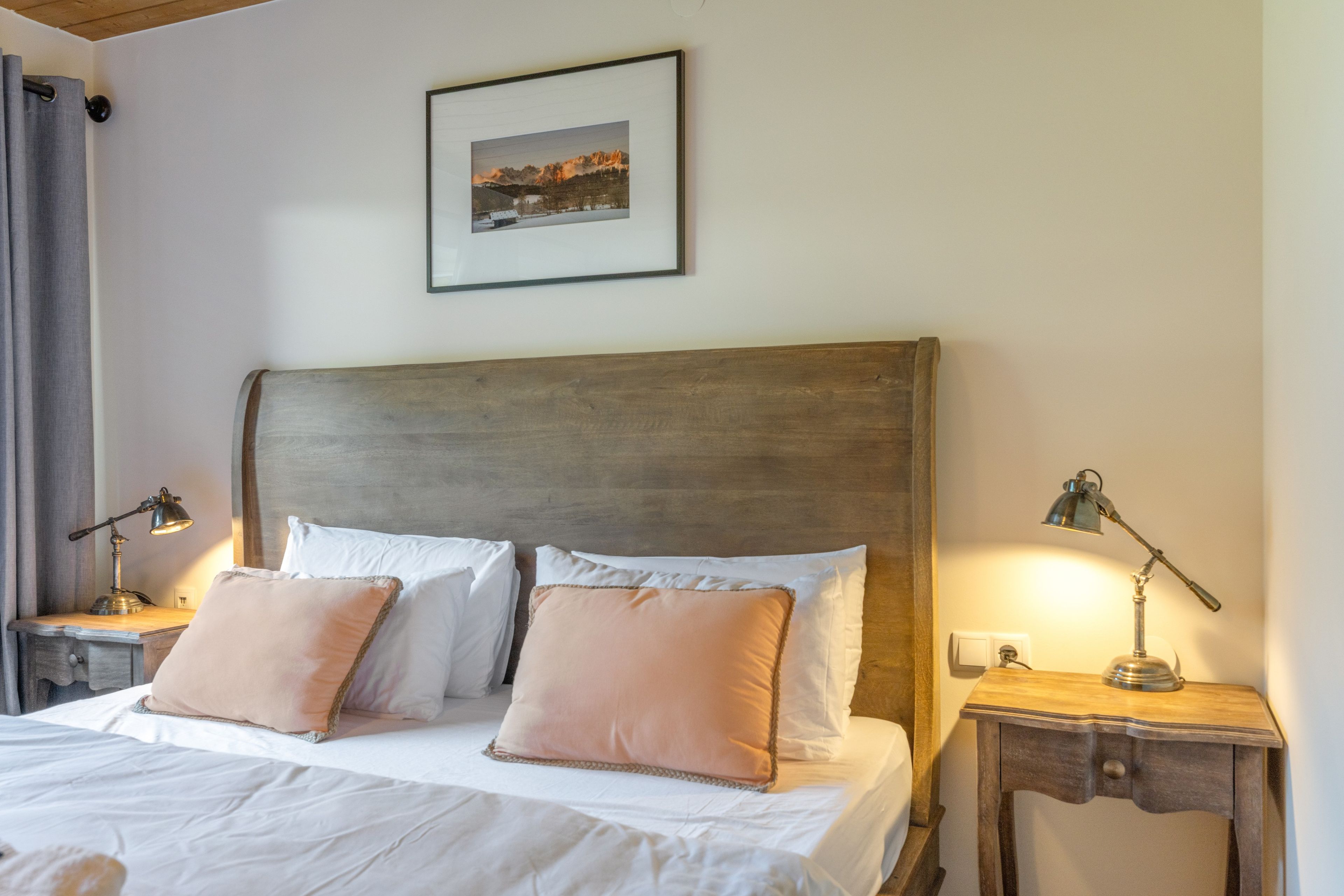 A neatly made bed with white linens and two pillows, a wooden headboard, a bedside table with a lamp, and a framed picture on the wall.