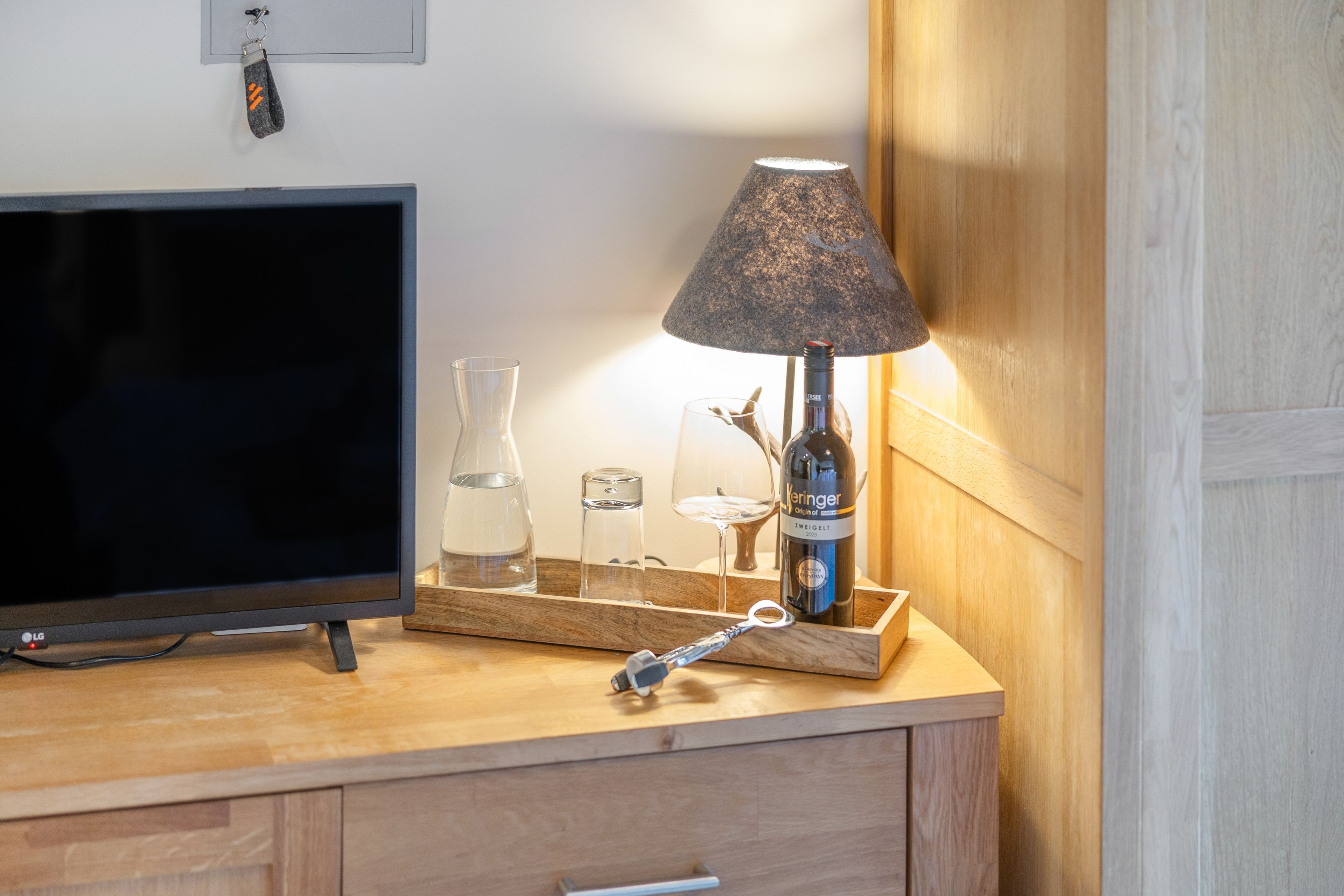 Un escritorio de madera con un televisor, una lámpara, una botella de vino y un abrelatas.