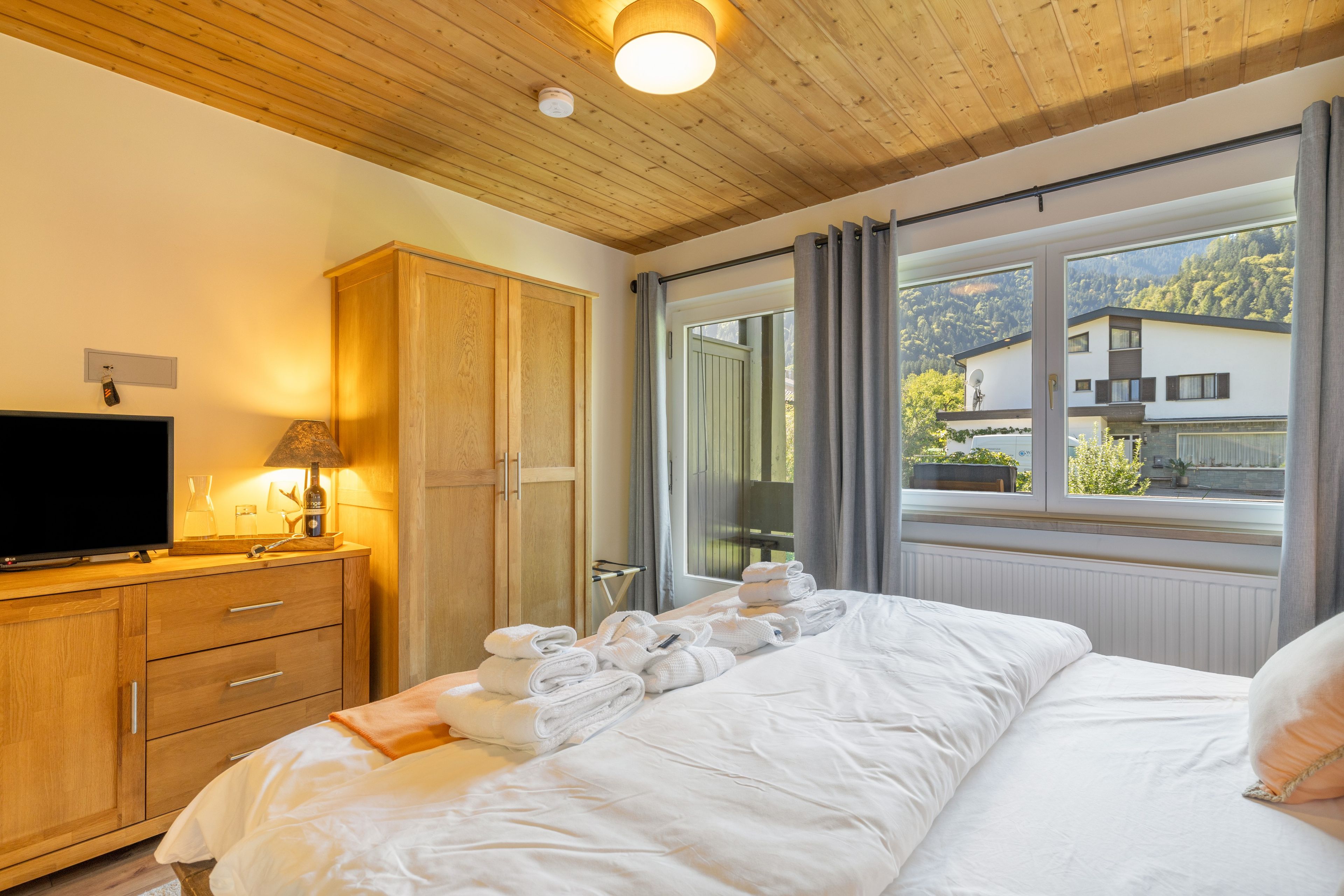 Ein Hotelzimmer mit Holzboden, einem Bett mit weißer Bettwäsche, einem Fernseher auf einer Holzkommode und Blick auf einen Berg durch ein Fenster.