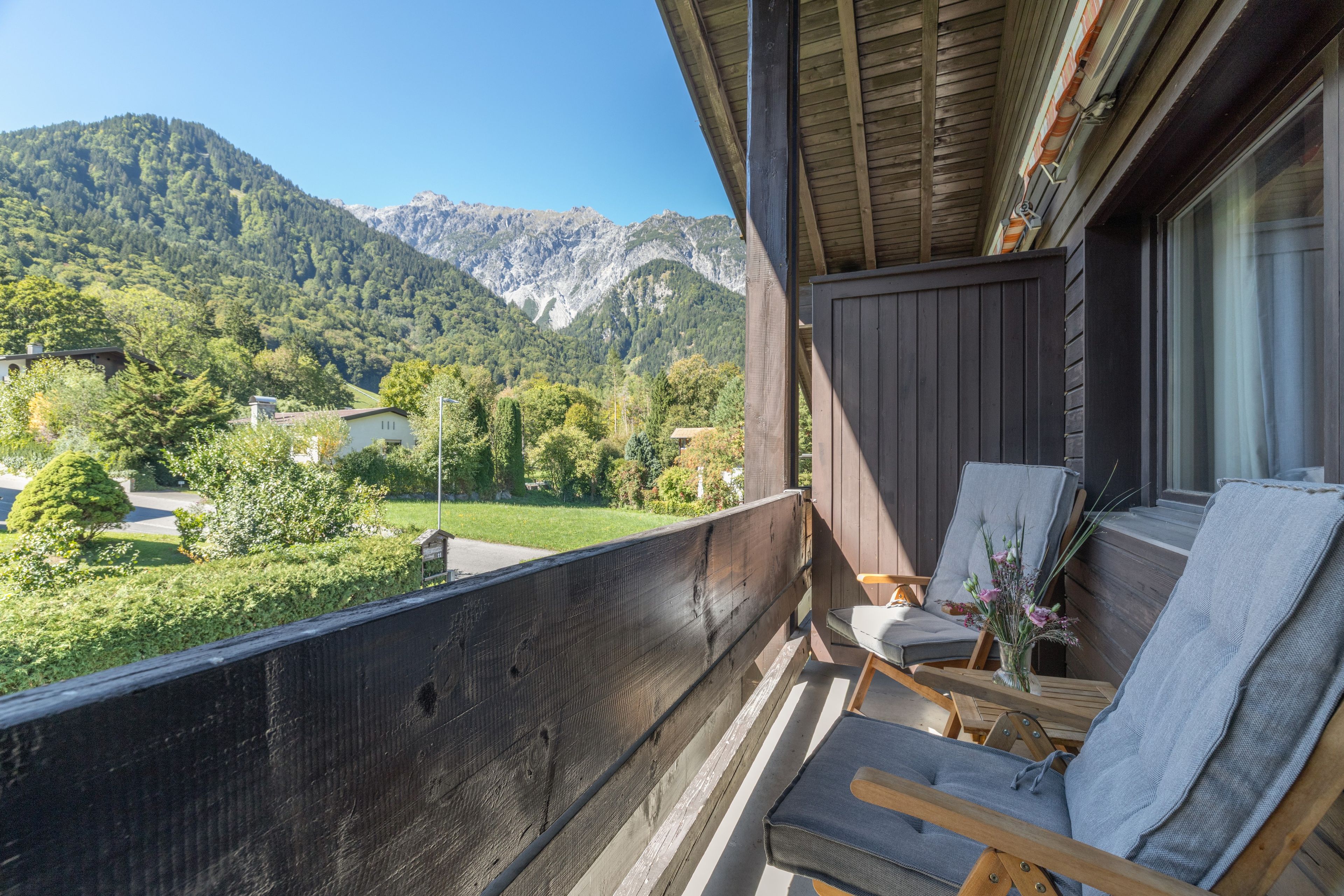 Ein Balkon mit Blick auf eine gebirgige Landschaft, Holzmöbel und ein kleiner Tisch mit einem Blumenstrauß.