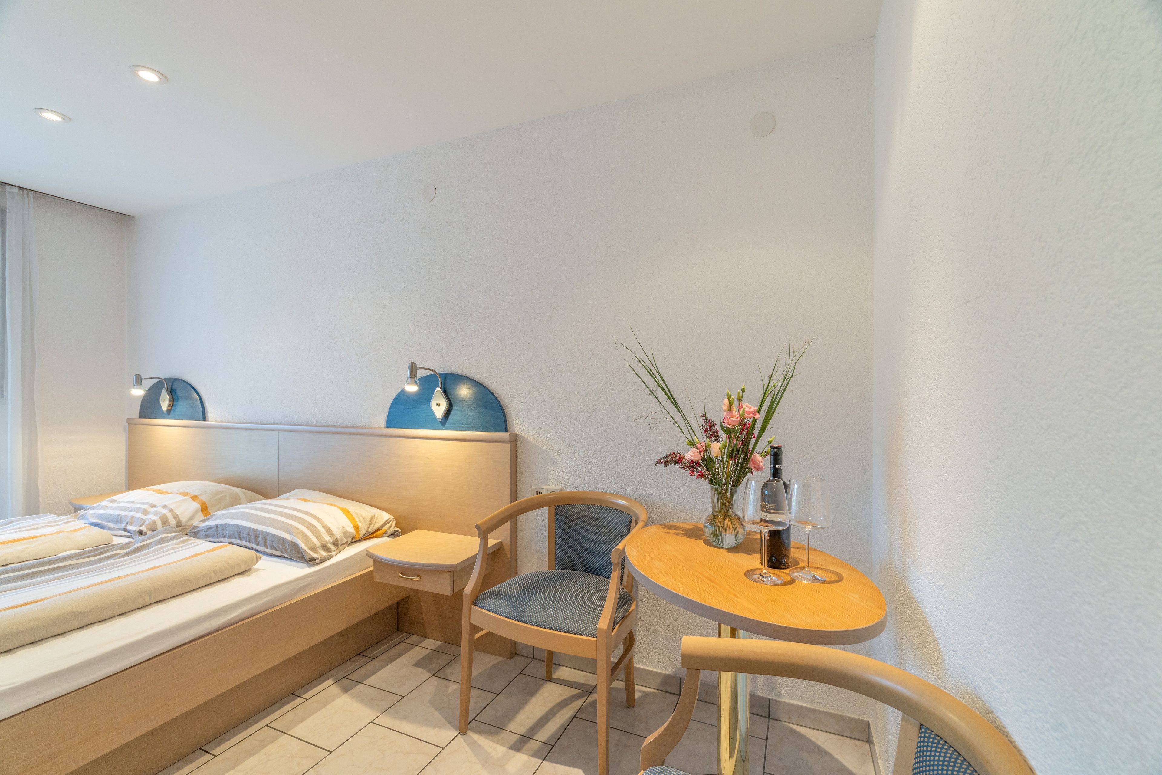 A hotel room with a bed, a small table with a vase of flowers, a chair, and a lamp.
