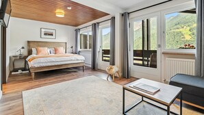 A bedroom with a wooden ceiling, a bed, a nightstand, a window with a view of the countryside, and a small table with a book. - Classic Double Room with Kitchenette, Terrace, and Mountain View (Vandans)