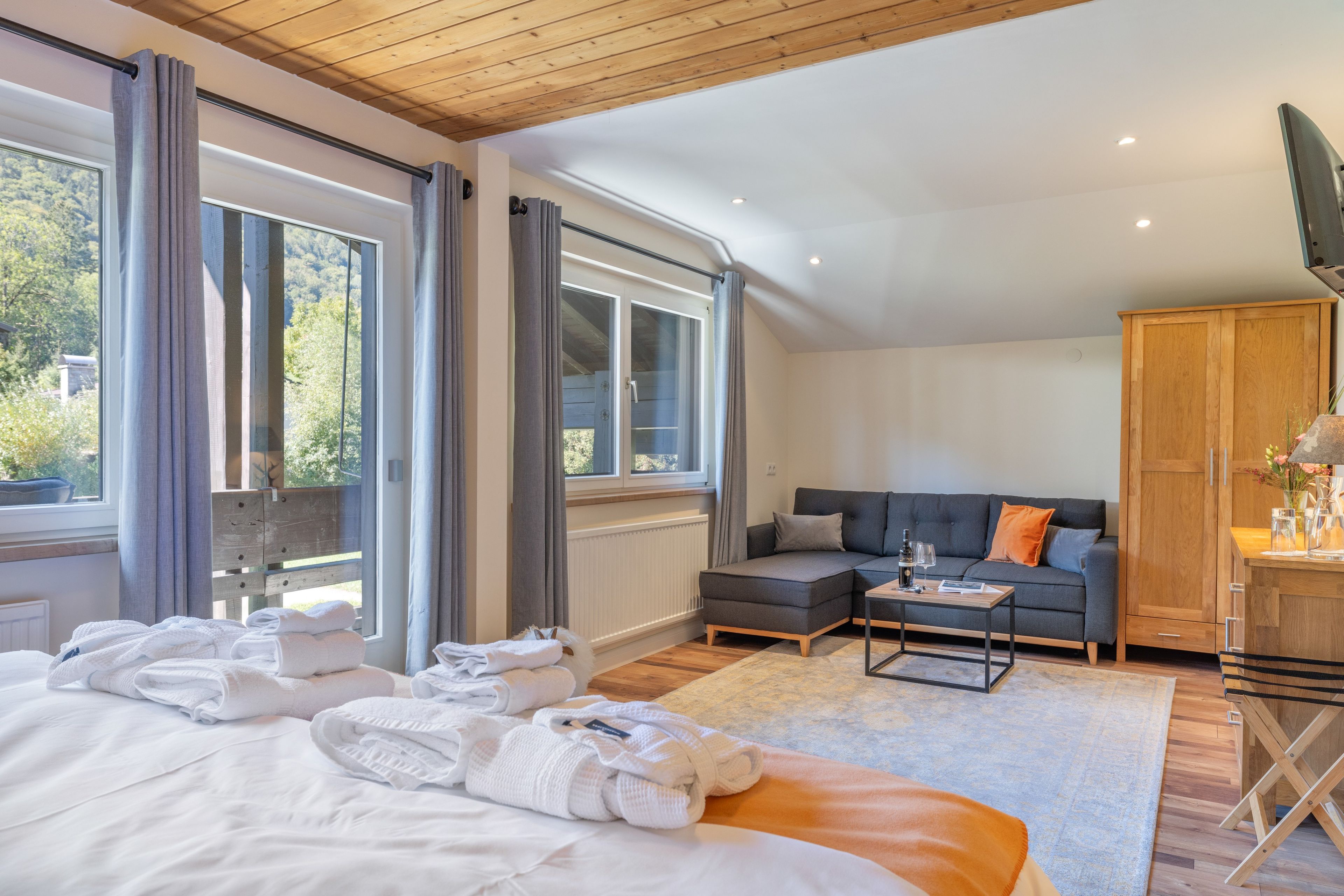 Ein modernes Schlafzimmer mit einem großen Bett, einer Couch und einem Esstisch. Es gibt Fenster mit Vorhängen, eine Holzbalkendecke und einen Blick auf Grünflächen außerhalb.
