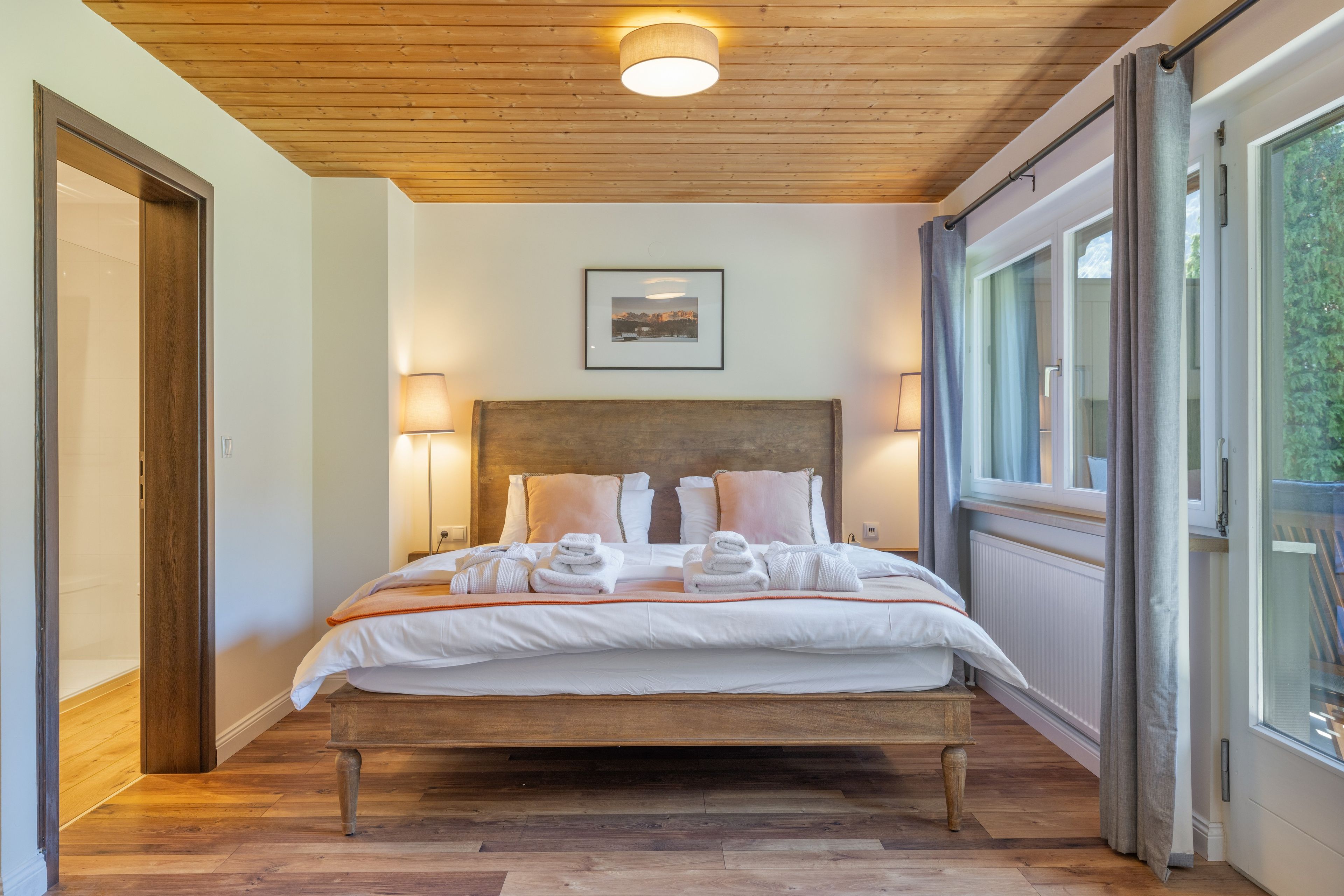 Ein Hotelzimmer mit Holzbalkendecke, einem Bett mit weißen Leinen und Handtüchern, einem gerahmten Bild an der Wand und einem Fenster mit Vorhängen.