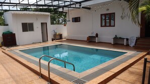 A swimming pool with a tiled deck, a ladder, and a covered area with string lights. - Tremendous 3-bedroom villa in pleasant Hosur getaway (Denkanikottai)
