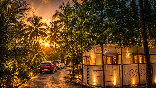 A paved pathway leading to a building with a glass facade, surrounded by palm trees and a sunset in the background. - Tremendous 3-bedroom villa in pleasant Hosur getaway (Denkanikottai)