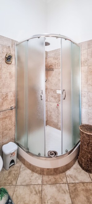 A glass-enclosed shower with a curved design, a small stool, and a wicker basket in a bathroom with beige tiles. - Magic DaLu House (Olbia)