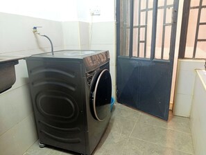 Eine kompakte, dunkel gefärbte Waschmaschine steht in einer Ecke eines Raumes mit Fliesenboden und einer Tür mit Gitterrost.