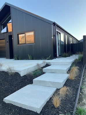 A modern building with a metal exterior, a concrete staircase leading to the entrance, and a small garden area with plants. - Modern, Cosy, Clean Apartment in Peaceful Area (Jack's Point)