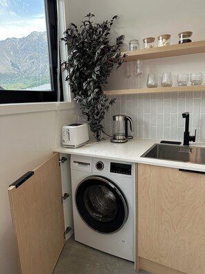 A compact kitchen with a washing machine, a toaster, a coffee maker, and a window with a view of mountains. - Modern, Cosy, Clean Apartment in Peaceful Area (Jack's Point)
