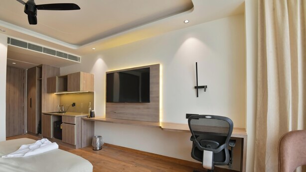 A hotel room with a bed, a desk with a chair, a kitchenette, and a ceiling fan. - Melange Hotel (Gurugram)