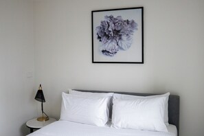 A neatly made bed with white linens and pillows, a black and gold lamp on a small bedside table, and a framed floral artwork on the wall. - Modern high rise apt, with balcony, secure parking (Southbank)