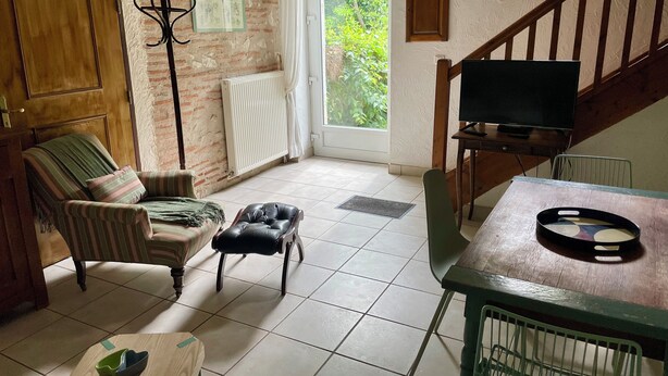 Una habitación con una escalera de madera, un televisor sobre una mesa de madera, un sillón a rayas, un sillón de cuero y un radiador.