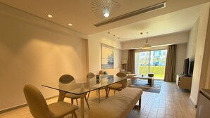 A modern living room with a glass dining table, beige sofas, and a large window offering a view of greenery. - Fouka North Coast SA-2B-B02 (Marsa Matrouh)