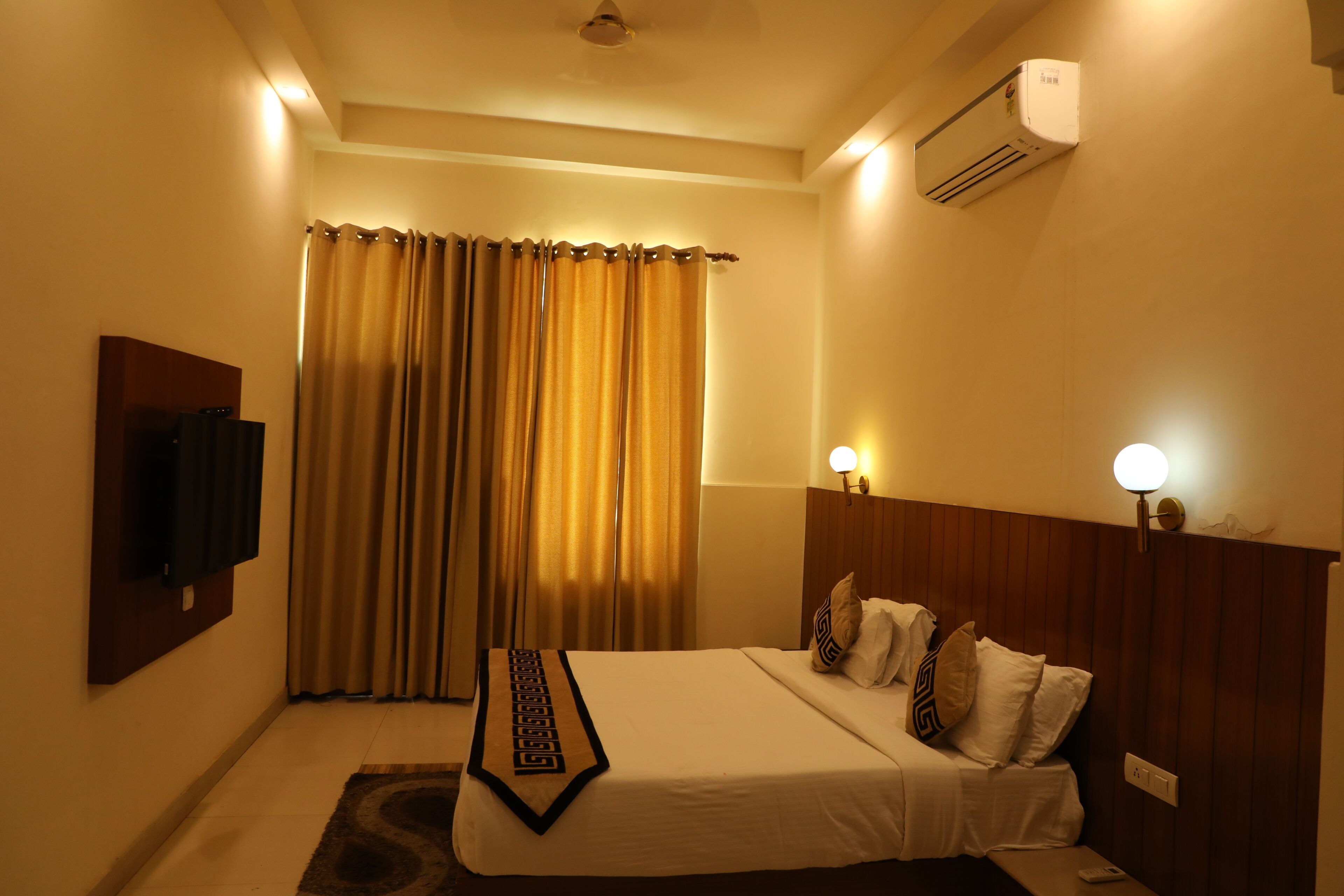 A hotel room with a bed, curtains, a flat-screen TV, a bedside table, and a wall-mounted air conditioner.