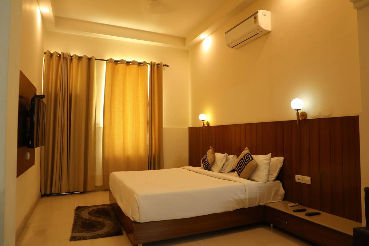 A hotel room with a bed, a bedside table, a wall-mounted TV, and an air conditioning unit.