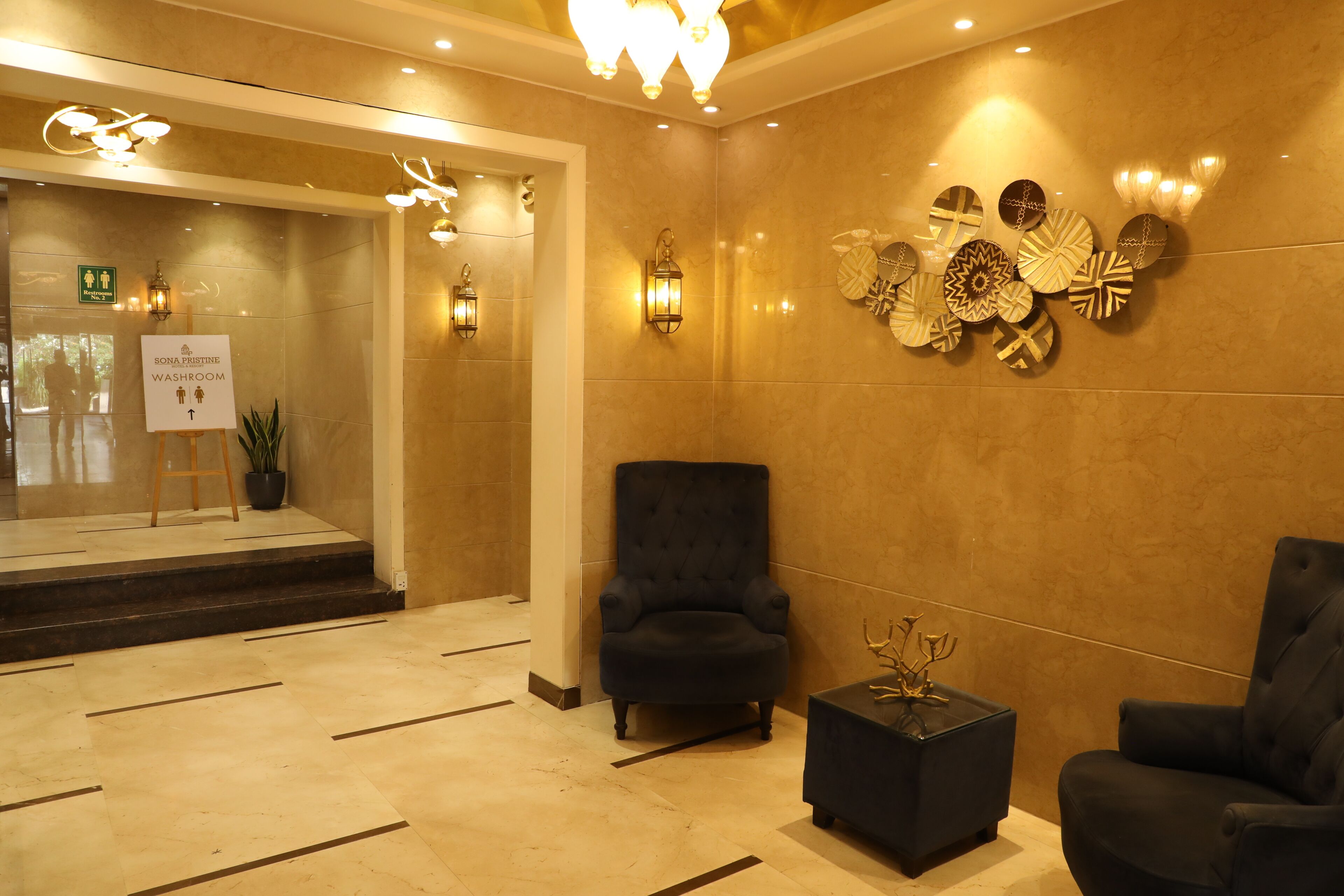A lobby with a black armchair, a small table with a decorative item, a wall-mounted light fixture, and a sign indicating a restroom.