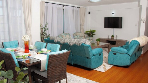Una sala de estar con sofás color turquesa, una mesa de comedor con sillas, un televisor montado en la pared y una pequeña planta en la mesa de centro.