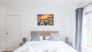 Une chambre d’hôtel avec un lit, deux tables de chevet, un tableau représentant des tournesols, une porte et une fenêtre avec des rideaux.