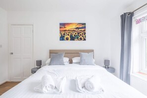 Ein Hotelzimmer mit einem Bett, zwei Nachttischen, einem Gemälde von Sonnenblumen, einer Tür und einem Fenster mit Vorhängen.