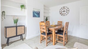 Un coin repas avec une table et des chaises en bois, une horloge au mur et une console en rotin agrémentée de plantes.