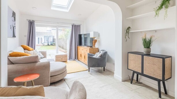 Un salon moderne avec un canapé, un pouf et une petite table. On y trouve également un téléviseur, un meuble et une fenêtre avec des rideaux.