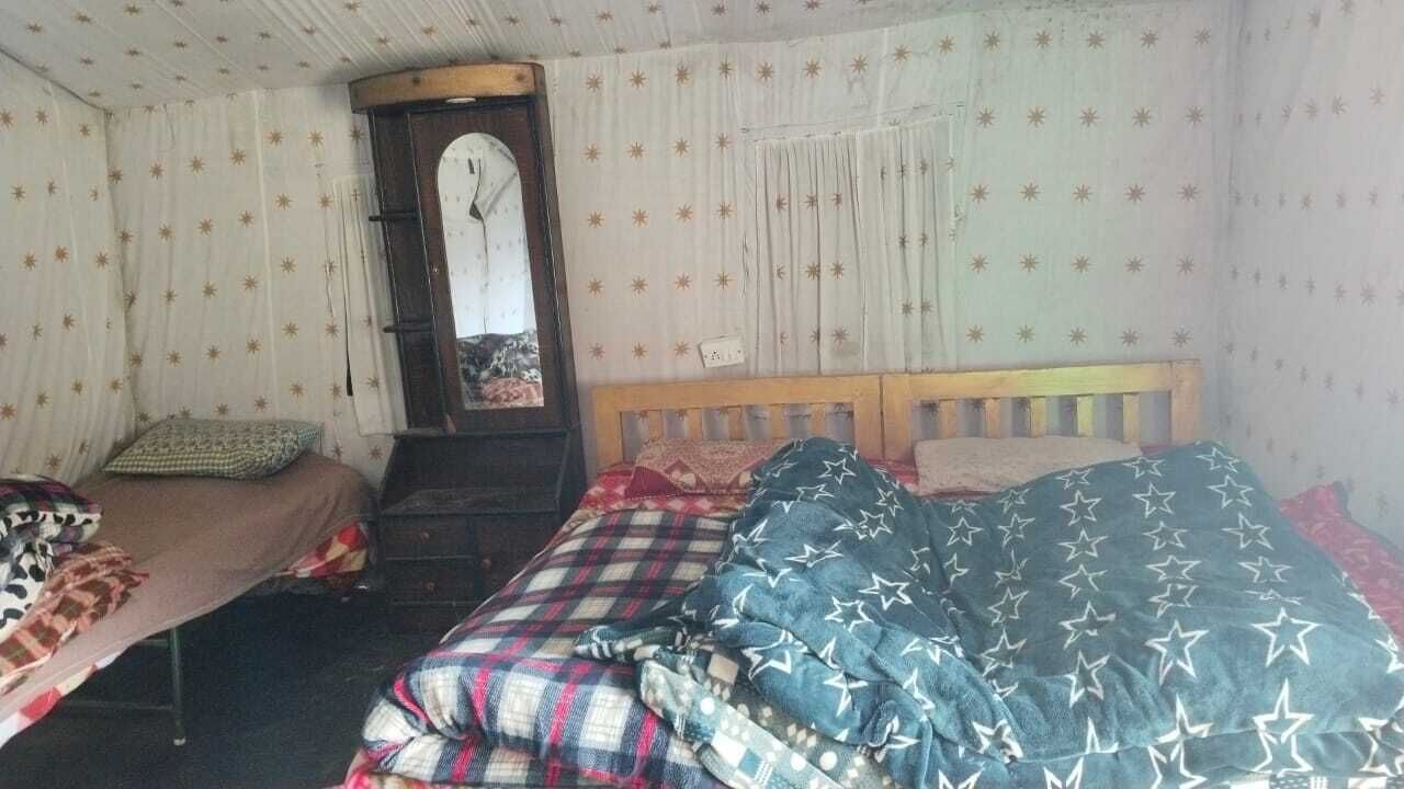 Una habitación con dos camas, una con cabecera de madera y la otra sin adornos. Hay un espejo en la pared, y el papel tapiz presenta un diseño decorativo.