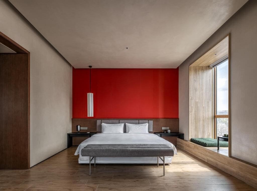 Quarto moderno com cama grande, parede de destaque vermelha e vista pela janela.