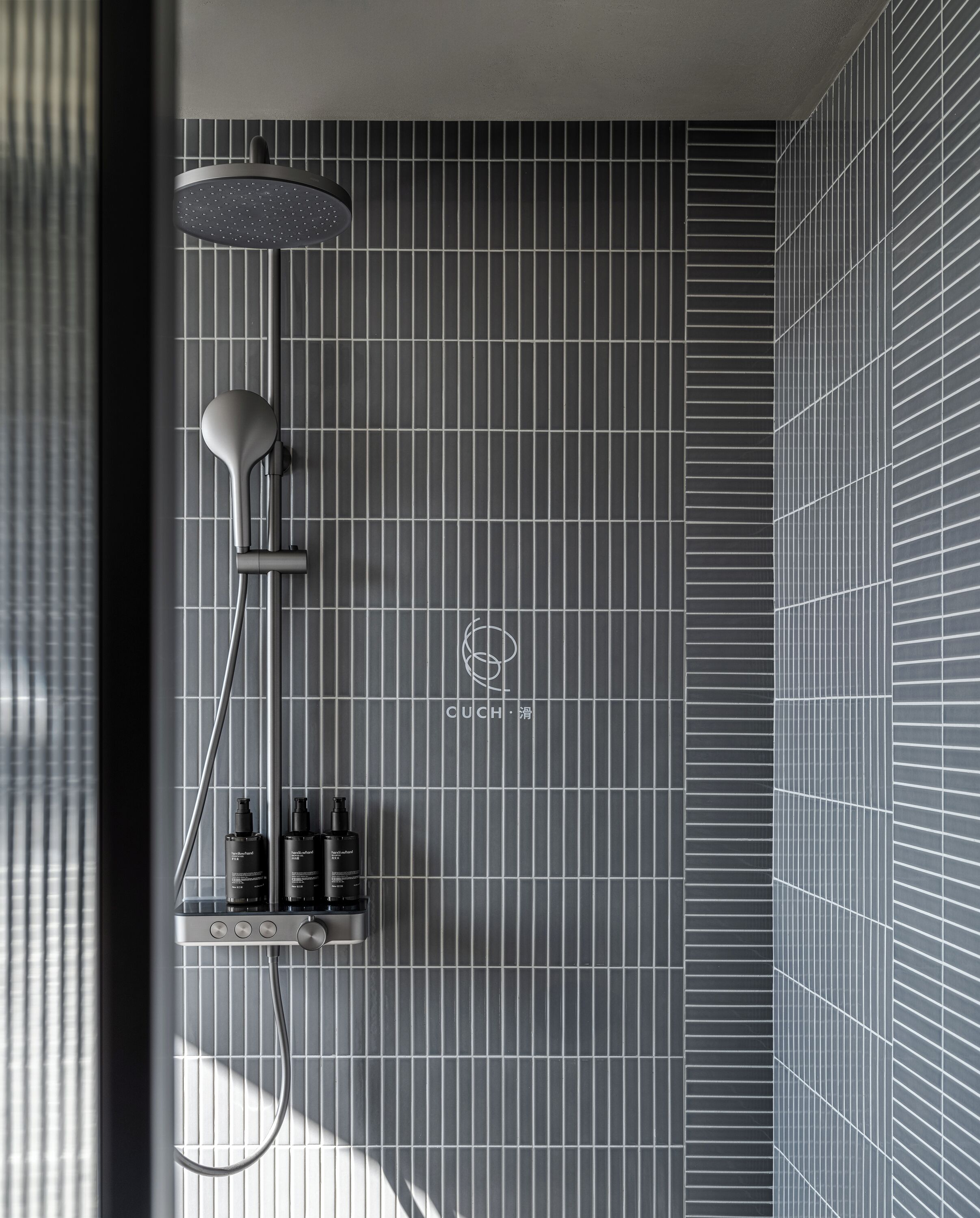 Une douche moderne dotée d’un pommeau de douche à pluie et d’un pommeau de douche à main, d’une étagère murale pour les articles de toilette et d’un mur carrelé avec un motif en réseau.