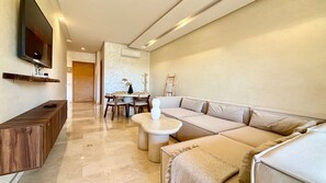 A modern living room with a sofa, a coffee table, a TV mounted on the wall, and a dining area with chairs and a table. - PRESTIGIA Saphir Marrakech View Pool (Marrakech)