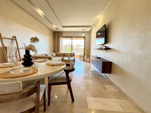 A modern dining area with a round table, chairs, a TV mounted on the wall, and a view of the outdoors. - PRESTIGIA Saphir Marrakech View Pool (Marrakech)