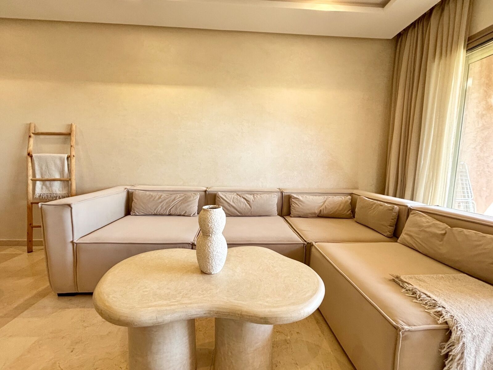 Un salon moderne avec un canapé d’angle beige, une table basse en pierre et une échelle en bois adossée au mur.